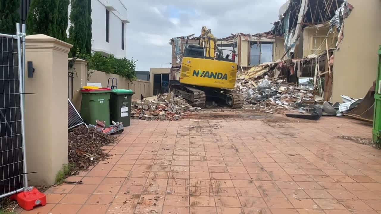 Excavator performing concrete demolition on a residential construction site, showing detailed concrete cross-section.